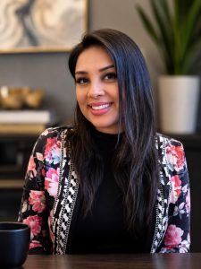 image of woman with dark brown hair and brown eyes wearing a black top with pink and black printed jacket in an office setting.